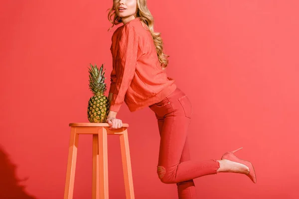 Cropped view of stylish woman posing with fresh pineapple at stool isolated on living coral — Stock Photo
