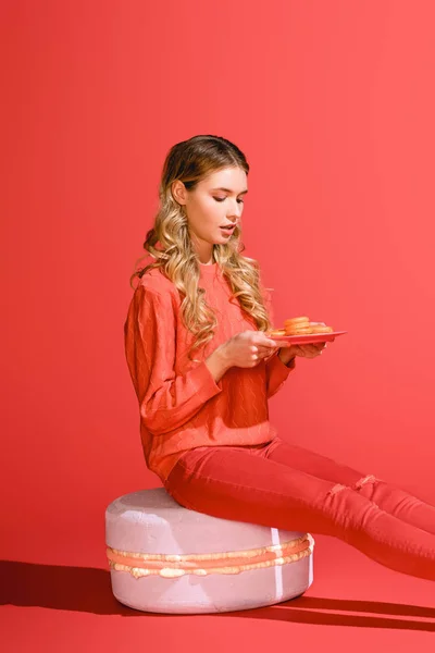 Beautiful girl holding plate with macaroons and sitting on big macaroon on living coral. Pantone color of the year 2019 concept — Stock Photo