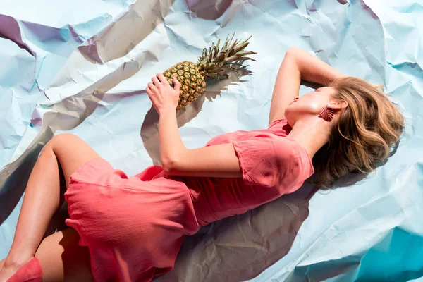 Draufsicht des eleganten Mädchens in lebendigem Korallenkleid, das Ananas hält und auf hellblauem zerknülltem Papier liegt — Stockfoto