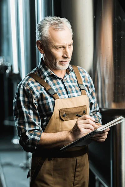 Профессиональный пивовар-мужчина в рабочем комбинезоне пишет в блокноте на пивоварне — стоковое фото