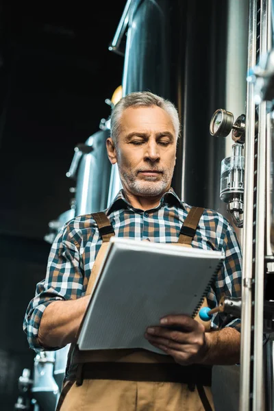 Cervecero senior serio escribiendo en bloc de notas mientras examina el equipo de la cervecería - foto de stock