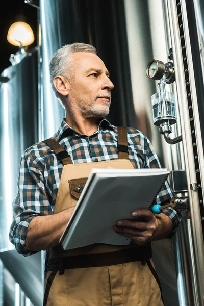 Schöner Brauer in Arbeitskleidung, der in Notizblock schreibt, während er die Brauereiausrüstung untersucht — Stockfoto