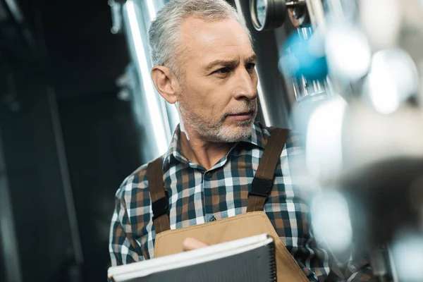 Brasseur senior écrit dans le bloc-notes tout en examinant l'équipement de brasserie — Photo de stock