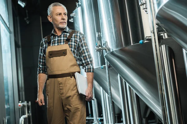 Cervecero senior en overoles de trabajo con bloc de notas mientras examina la cervecería - foto de stock