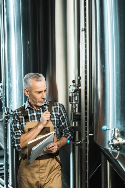 Guapo cervecero senior en overoles de trabajo sosteniendo bloc de notas y mirando el equipo de la cervecería - foto de stock