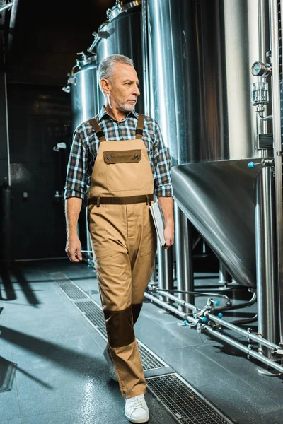 Cervejeiro sênior em macacões de trabalho segurando bloco de notas enquanto caminha e examina cervejaria — Fotografia de Stock