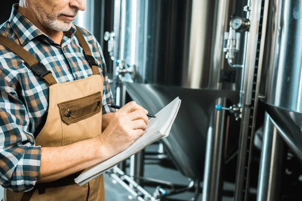 Vista recortada de cervecero en overoles de trabajo escrito en bloc de notas mientras se examina el equipo de la cervecería - foto de stock