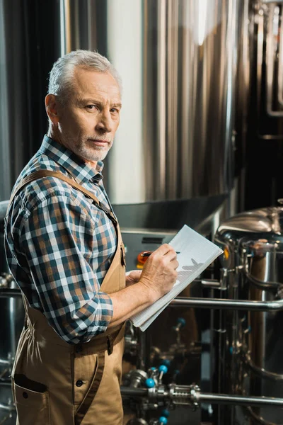 Senior brewer writing in notepad while examining brewery equipment — Stock Photo