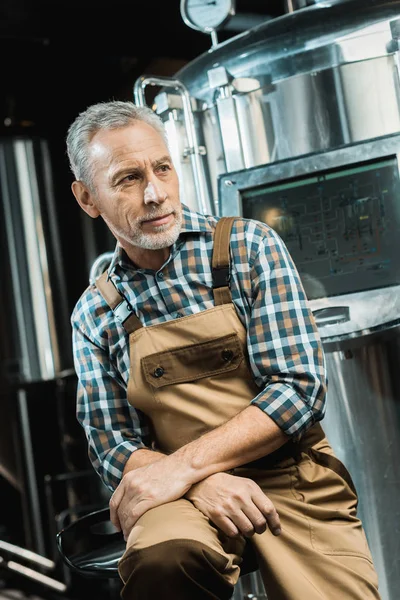 Senior male brewer in working overalls sitting near brewery equipment with screen — Stock Photo