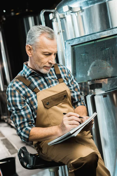Senior brewer in working overalls writing in notepad while examining brewery equipment — Stock Photo