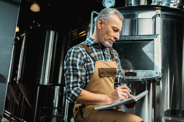 Professioneller Brauer in Arbeitskleidung, der in Notizblock schreibt, während er die Brauereiausrüstung untersucht — Stockfoto