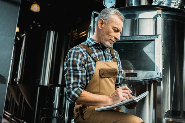 Brasseur professionnel en salopette de travail écriture dans le bloc-notes tout en examinant l'équipement de brasserie — Photo de stock