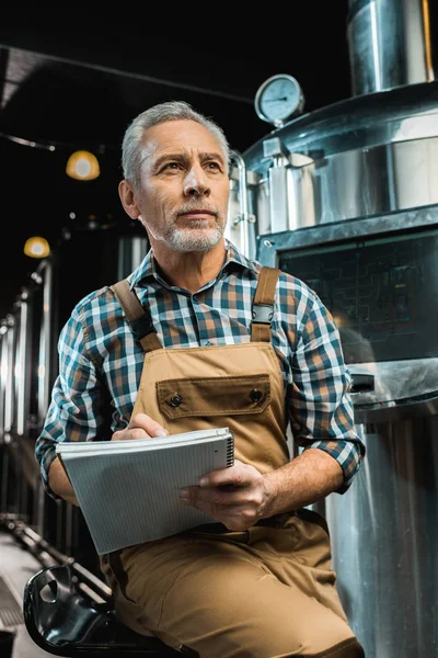 Thoughtful brewer in working overalls writing in notepad near brewery equipment — Stock Photo