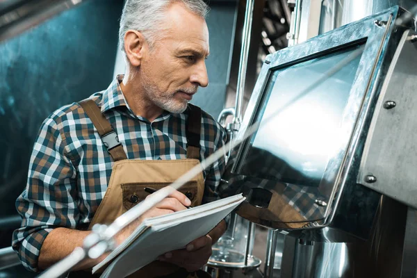 Senior brewer writing in notepad while looking at screen of brewery equipment — Stock Photo