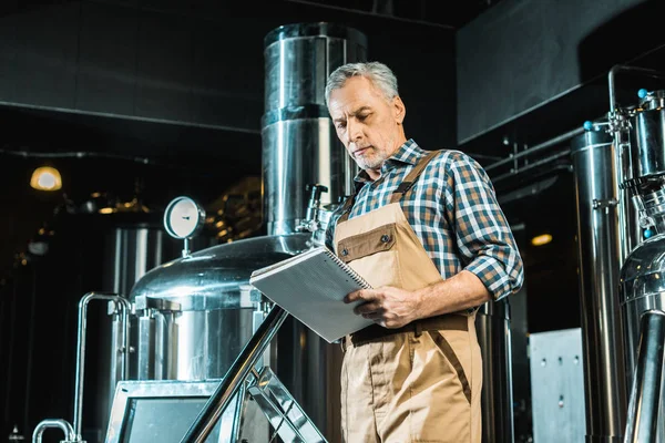 Braumeister in Arbeitskleidung schaut auf Notizblock, während er die Brauereiausrüstung untersucht — Stockfoto