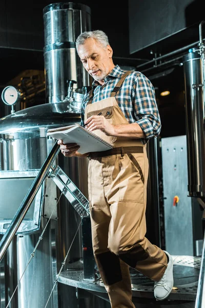 Handsome brewer in working overalls writing in notepad while examining brewery — Stock Photo
