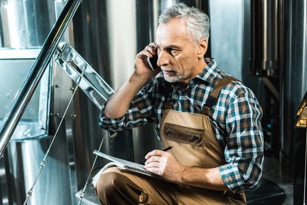 Handsome senior brewer in working overalls talking on smartphone while writing in notepad in brewery — Stock Photo