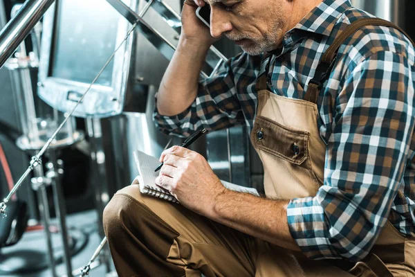 Senior brewer in working overalls talking on smartphone while writing in notepad in brewery — Stock Photo