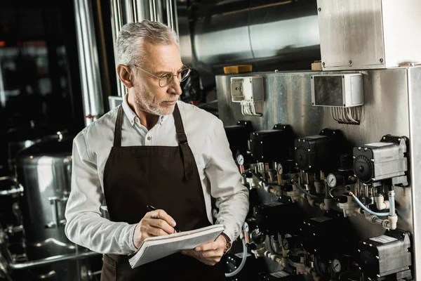 Guapo cervecero senior escribiendo en bloc de notas mientras revisa el equipo de la cervecería - foto de stock