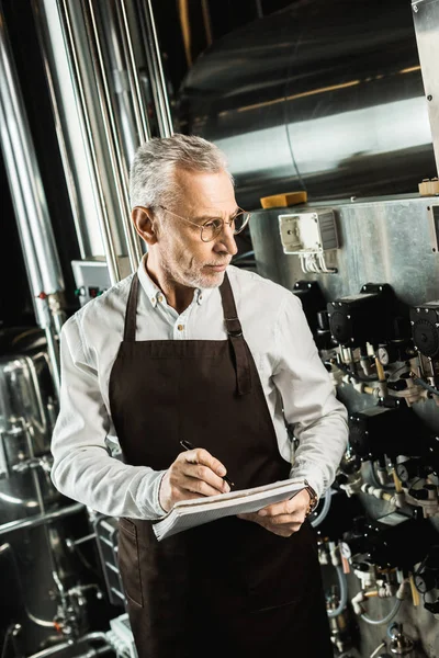 Senior-Brauer schreibt in Notizblock, während er Brauereiausrüstung untersucht — Stockfoto