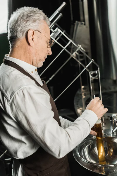 Professional senior brewer examining beer in flask in brewery — Stock Photo