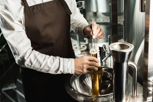 Vista recortada de cervecero profesional examinando cerveza en frasco en cervecería - foto de stock