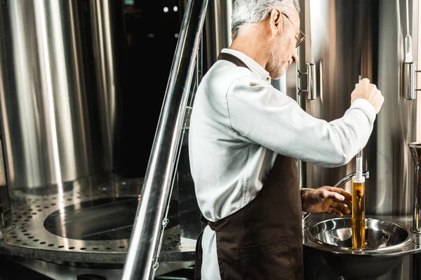 Uomo anziano con capelli grigi che esamina birra in fiaschetta in birreria — Foto stock