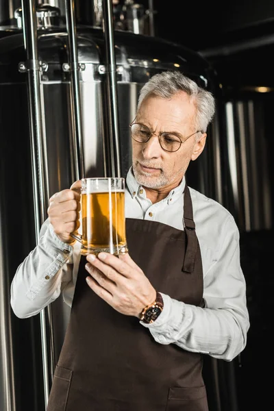Cervecero senior serio mirando un vaso de cerveza en la cervecería - foto de stock