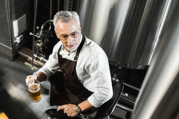 Vue aérienne du brasseur senior dans le tablier assis sur la chaise et tenant un verre de bière dans la brasserie — Photo de stock