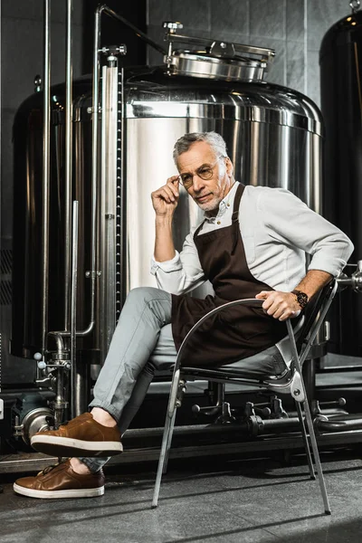 Senior brewer in apron sitting on chair in brewery — Stock Photo