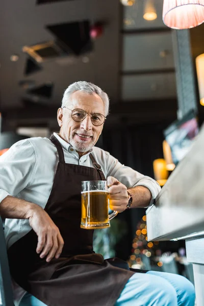 Senior worker in apron holding glass of beer near bar counter — Stock Photo