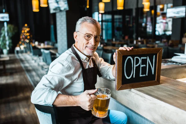 Smiling owner of pub holding open sign and glass of beer at bar counter — Stock Photo