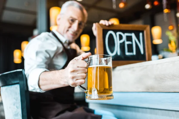 Selective focus of senior owner of pub holding open sign and glass of beer — Stock Photo