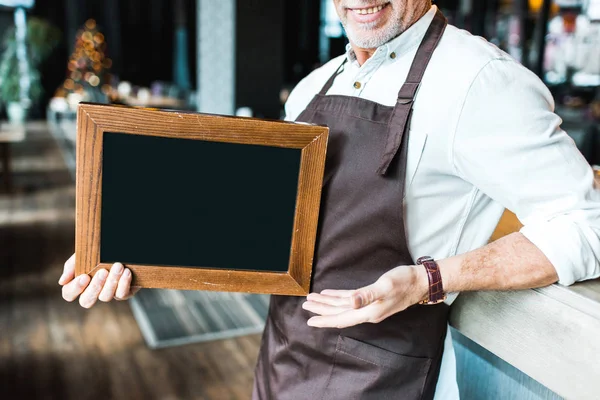 Vista ritagliata del proprietario senior di pub in possesso di bordo vuoto e in piedi vicino al bancone del bar — Foto stock