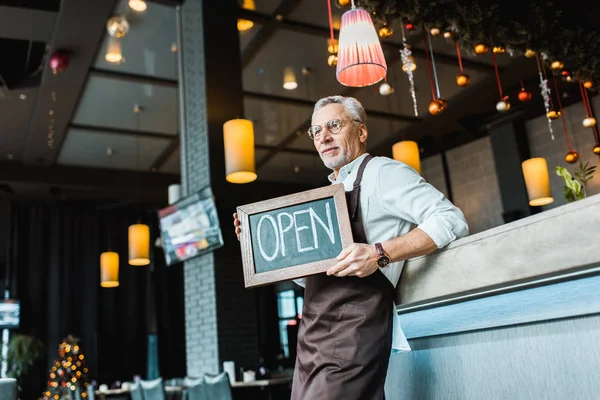 Senior worker of pub holding open sign and standing near bar counter — Stock Photo