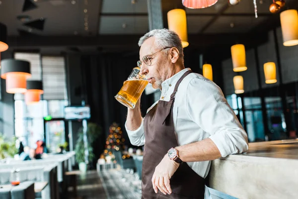 Senior male worker in apron holding drinking beer in pub — Stock Photo