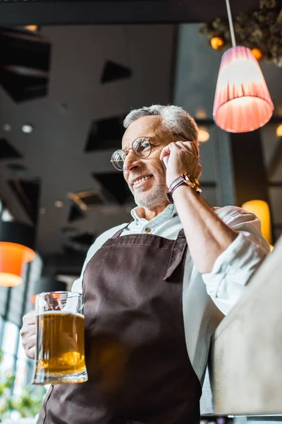 Senior man in apron talking on smartphone and holding glass of beer in pub — Stock Photo