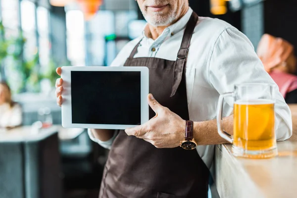Vista recortada del propietario masculino sosteniendo tableta digital con pantalla en blanco en el pub con vaso de cerveza - foto de stock