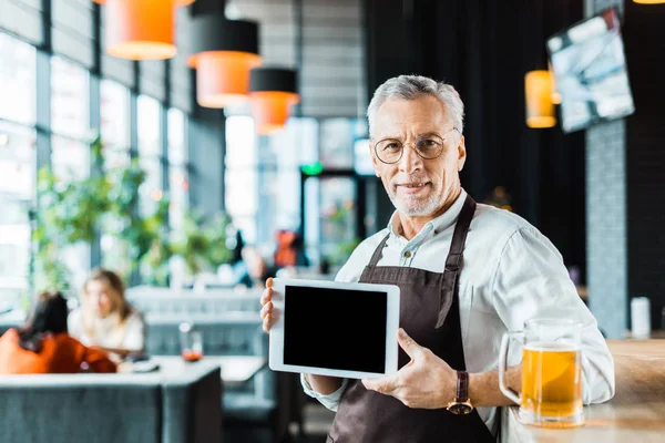 Propietario senior en delantal sosteniendo tableta digital con pantalla en blanco en pub con vaso de cerveza - foto de stock