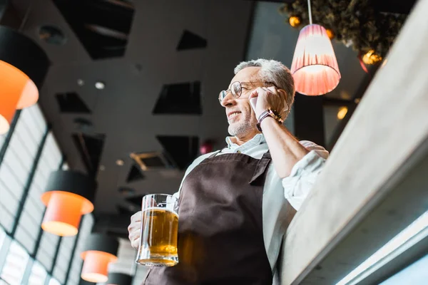 Blick von unten auf einen älteren Mann in Schürze, der mit dem Smartphone spricht und in der Kneipe ein Glas Bier hält — Stockfoto