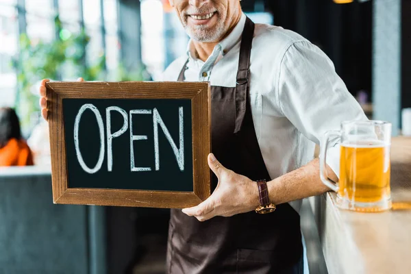 Cropped view of owner of pub holding open sign and standing near bar counter with glass of beer — Stock Photo