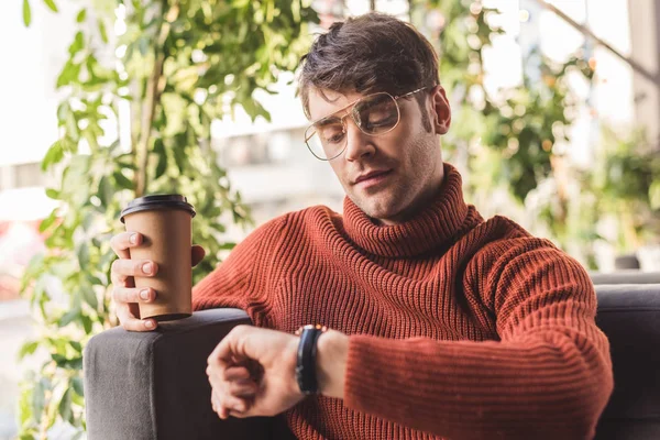 Pensive man in glasses holding paper cup while looking at watch in cafe — Stock Photo