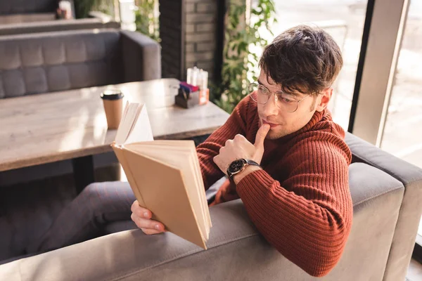 Hombre pensativo en gafas libro de lectura cerca de taza de papel en la cafetería - foto de stock