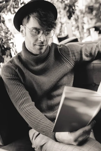 Hombre guapo en gafas y libro de lectura de sombrero mientras está sentado en la cafetería, fotografía en blanco y negro - foto de stock