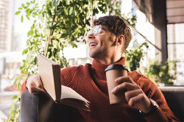 Joven alegre en gafas sosteniendo libro y taza de papel en las manos - foto de stock