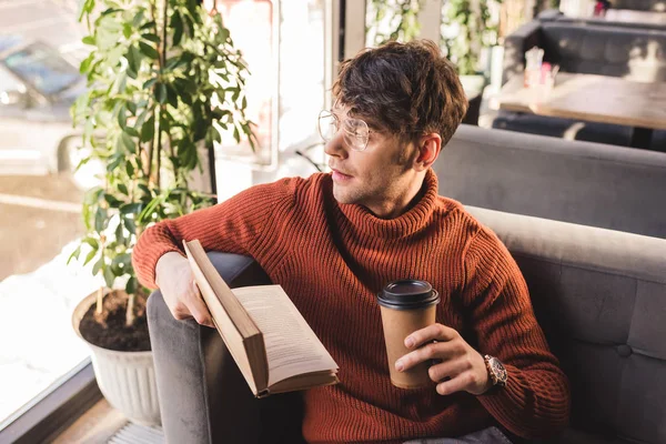 Joven pensativo en gafas sosteniendo libro y taza de papel en las manos - foto de stock