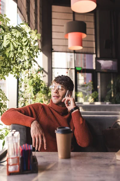 Селективный фокус красивого мужчины в очках, разговаривающего на смартфоне возле бумажной чашки с кофе — стоковое фото