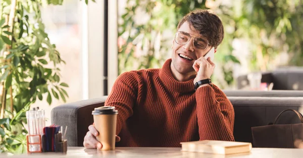 Messa a fuoco selettiva di uomo allegro in occhiali che parla su smartphone vicino a tazza di carta con caffè e libro — Foto stock