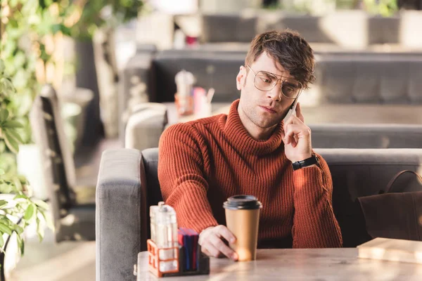 Enfoque selectivo de hombre serio en gafas hablando en el teléfono inteligente cerca de la taza de papel con café y libro - foto de stock