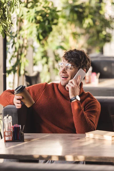 Enfoque selectivo de hombre feliz en gafas hablando en el teléfono inteligente y sosteniendo la taza de papel con café cerca del libro - foto de stock